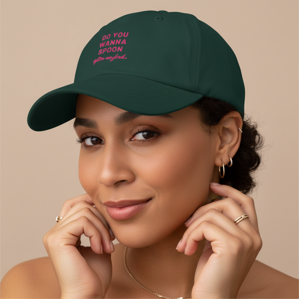 Dark green dad hat with "Do You Wanna Spoon After We Fork" in pink embroidery, worn by a queer femme with brown skin and curls.