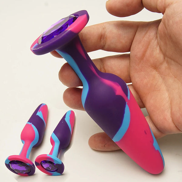 Hand holding a purple and pink jeweled butt plug with a flared crystal base; two matching plugs lie below