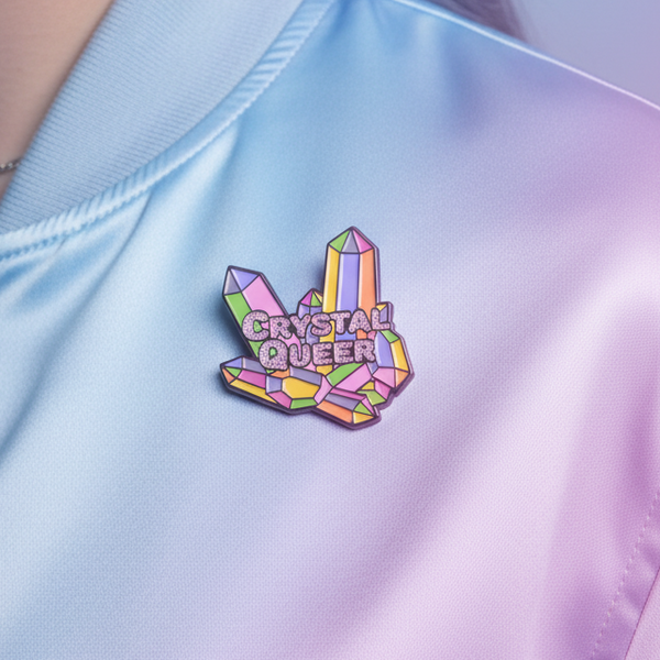 Close-up of a crystal-shaped enamel pin reading “CRYSTAL QUEER” on pastel fabric