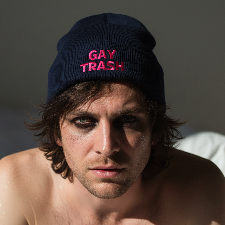 Gay Trash Cuffed Beanie with "GAY TRASH" in pink, worn by a queer person with tousled brown hair and glittery makeup.