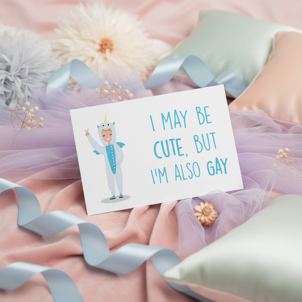 Postcard showing a cartoon human in a unicorn onesie, with “I MAY BE CUTE, BUT IM ALSO GAY” and blue ribbon, tulle