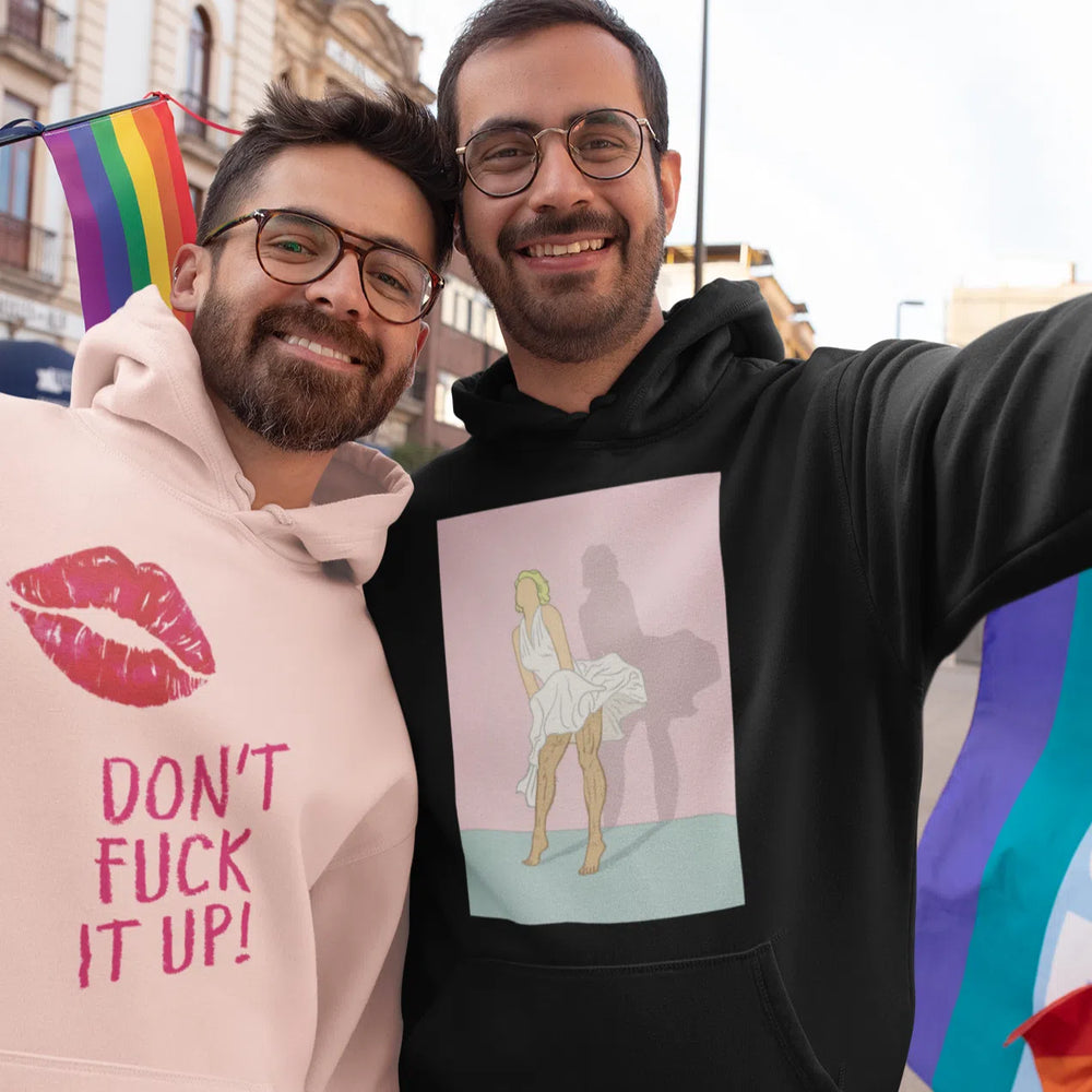Two people wearing graphic hoodies beside rainbow Pride flags; black hoodie shows a seated figure illustration
