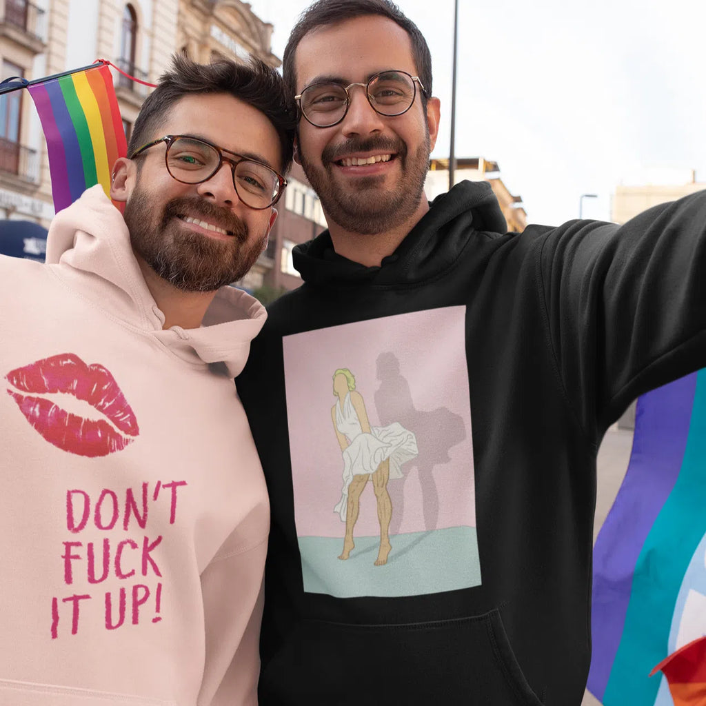 Two people wearing graphic hoodies beside rainbow Pride flags; black hoodie shows a seated figure illustration
