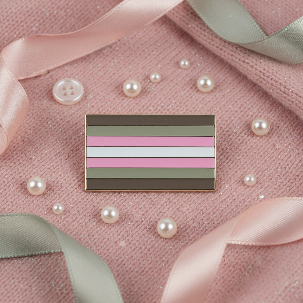 Demigirl pride enamel pin with horizontal brown, gray, pink, and white stripes on pink fabric