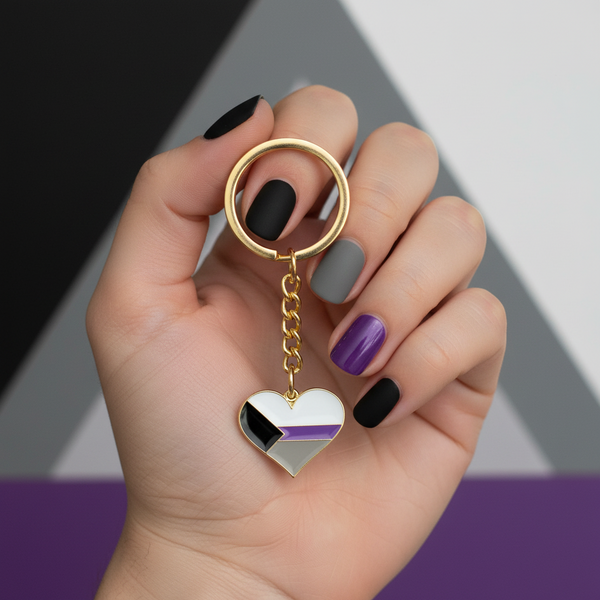 Hand holding a gold demisexual pride heart keychain with black, gray, white, and purple stripes