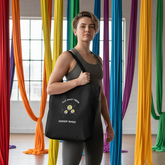 Fly Away From Gender Roles Eco Tote Bag in black canvas with planet motifs and empowering slogan, slung over shoulder in aerial-yoga studio, embodying prideful, queer visibility and sustainable self-expression.