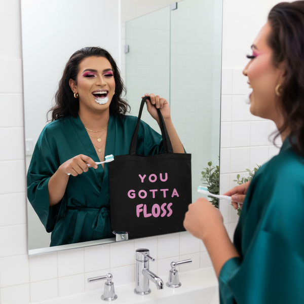 Person proudly holds up a YOU GOTTA FLOSS Eco Tote Bag in a bathroom, embodying queer pride and self-expression. The organic cotton tote is perfect for eco-conscious, bold, and joyful individuals.
