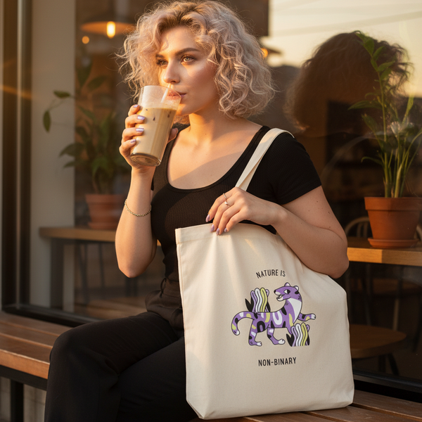 Person sipping iced coffee while displaying a cream tote bag reading “NATURE IS NON-BINARY”