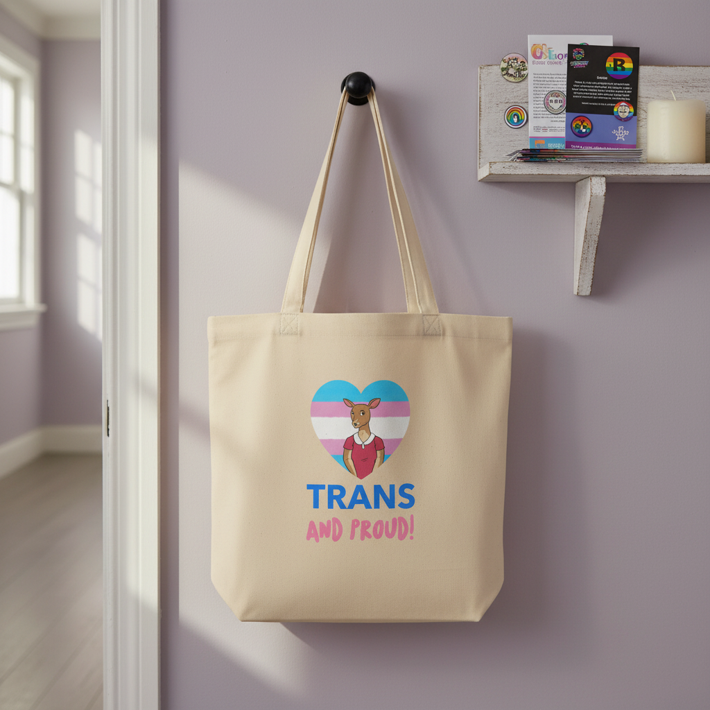 Beige organic cotton "Trans And Proud" tote bag with a cartoon bear and trans flag heart, on a black hook by pins and cards.