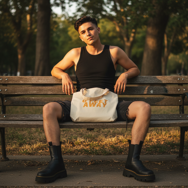 Young person on a park bench at golden hour with a Zero Feet Away Grindr Eco Tote Bag in bold yellow letters, showcasing queer empowerment and eco-conscious style with prideful self-expression.