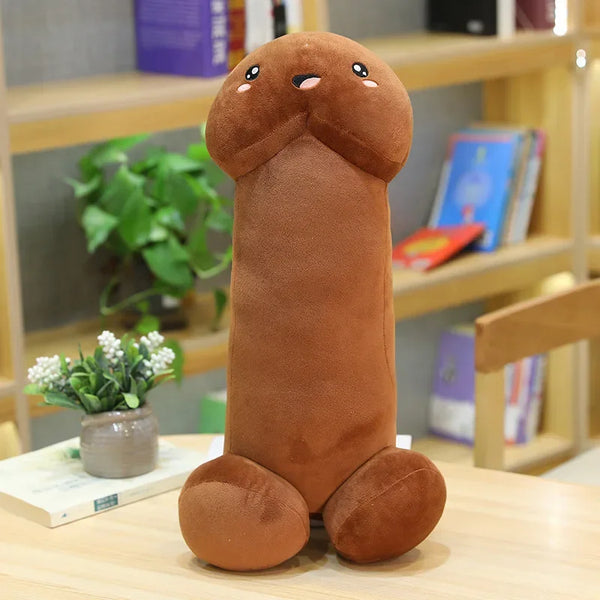 Brown penis-shaped plush pillow with a smiling face and rounded base, standing on a table