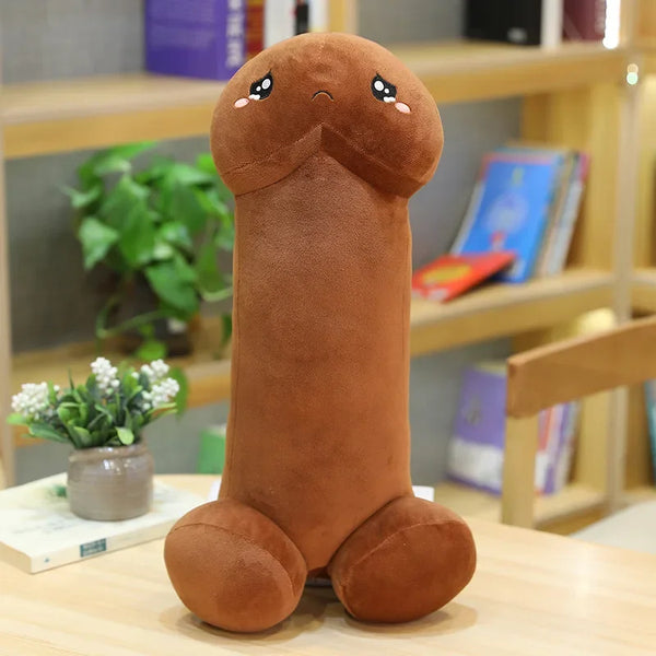 Brown penis-shaped plush pillow with a sad embroidered face, seated on a table