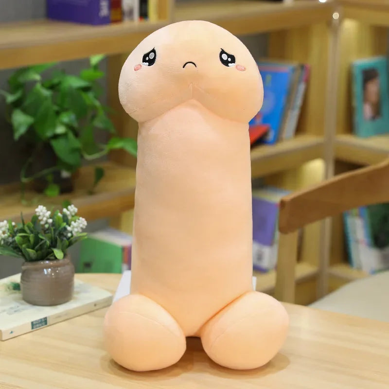 Peach-colored penis-shaped plush pillow with a shy embroidered face, seated on a table