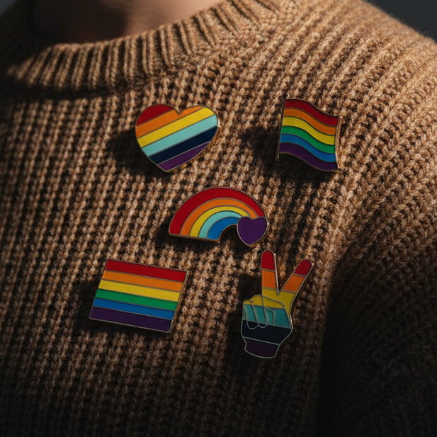 Five rainbow Pride enamel pins on a brown knit sweater, including hearts, a rainbow, and peace sign