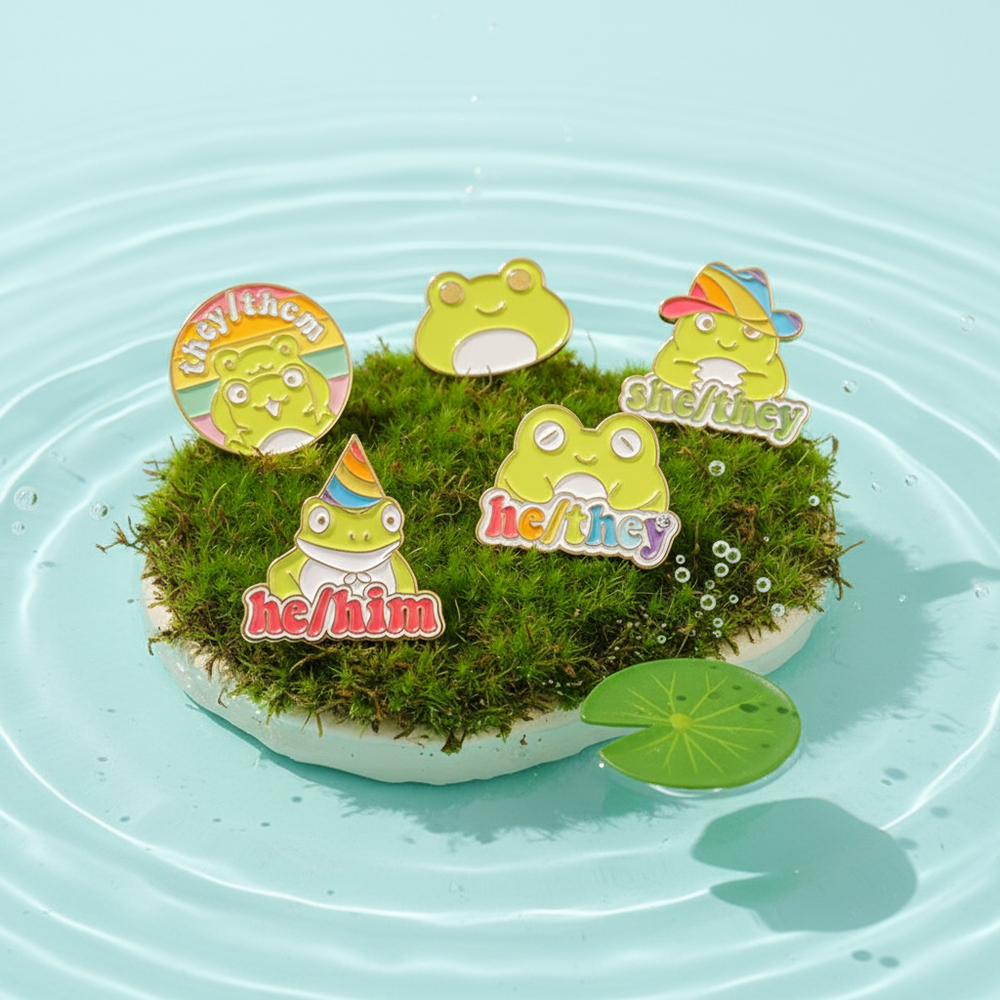 Four frog enamel pins reading “any pronouns,” “he/him,” “he/they,” and “she/they” on moss in water