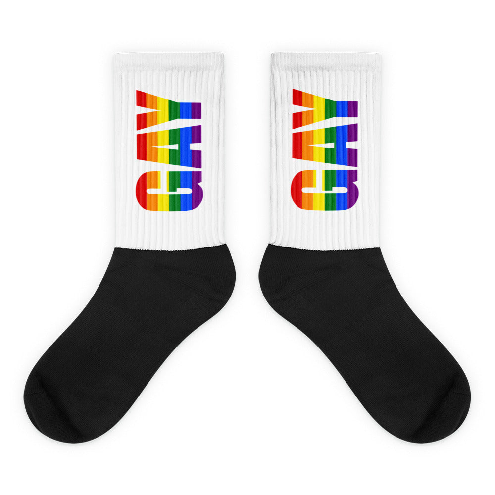 Pair of crew socks with black feet and white ribbed cuffs reading GAY in rainbow stripes
