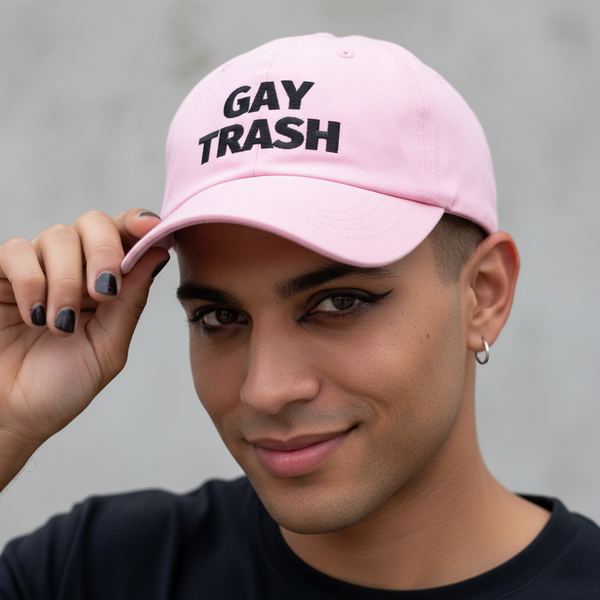 Gay Trash (Black Text) Cap—a pink hat with bold black lettering—tipped by a smiling, short-haired queer with winged eyeliner.