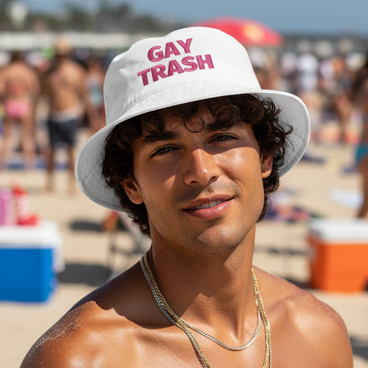 Gay Trash Bucket Hat in white cotton with bold pink "GAY TRASH" lettering, worn by a shirtless person at a busy, sunny beach