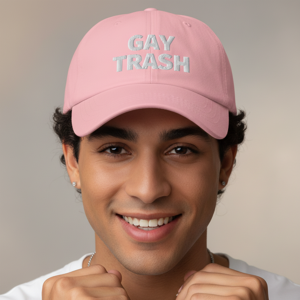 Light pink embroidered dad hat reads "GAY TRASH" in bold white letters; person with short curls, hoop earrings, smiling