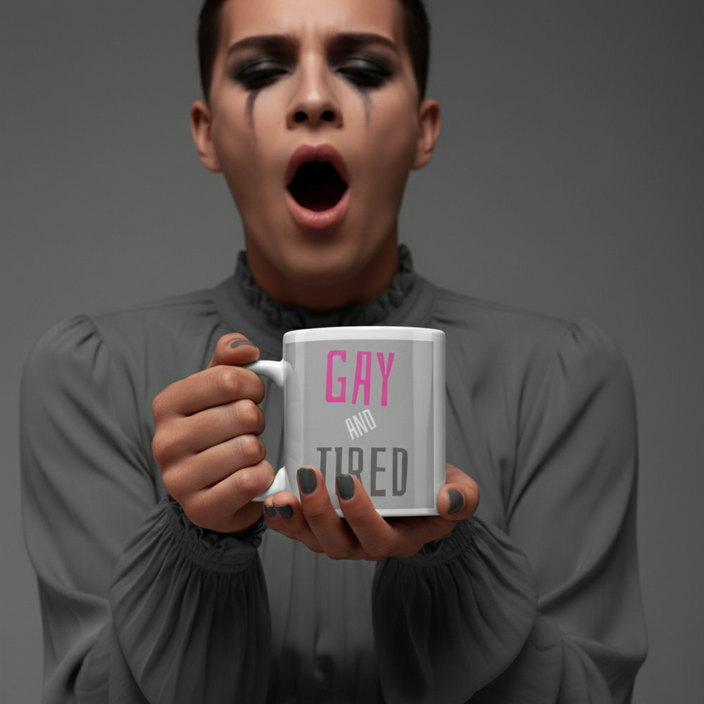 Person with dramatic eye makeup holds a white mug reading “GAY AND TIRED” while yawning