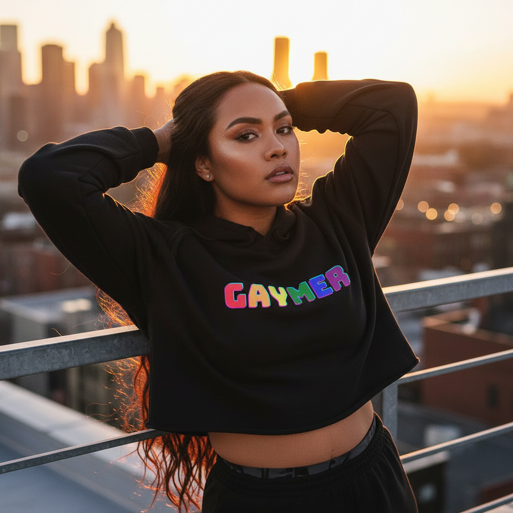 Gaymer Crop Hoodie with bold rainbow "GAYMER" lettering, worn by a person at sunset on a city rooftop, golden skyline behind