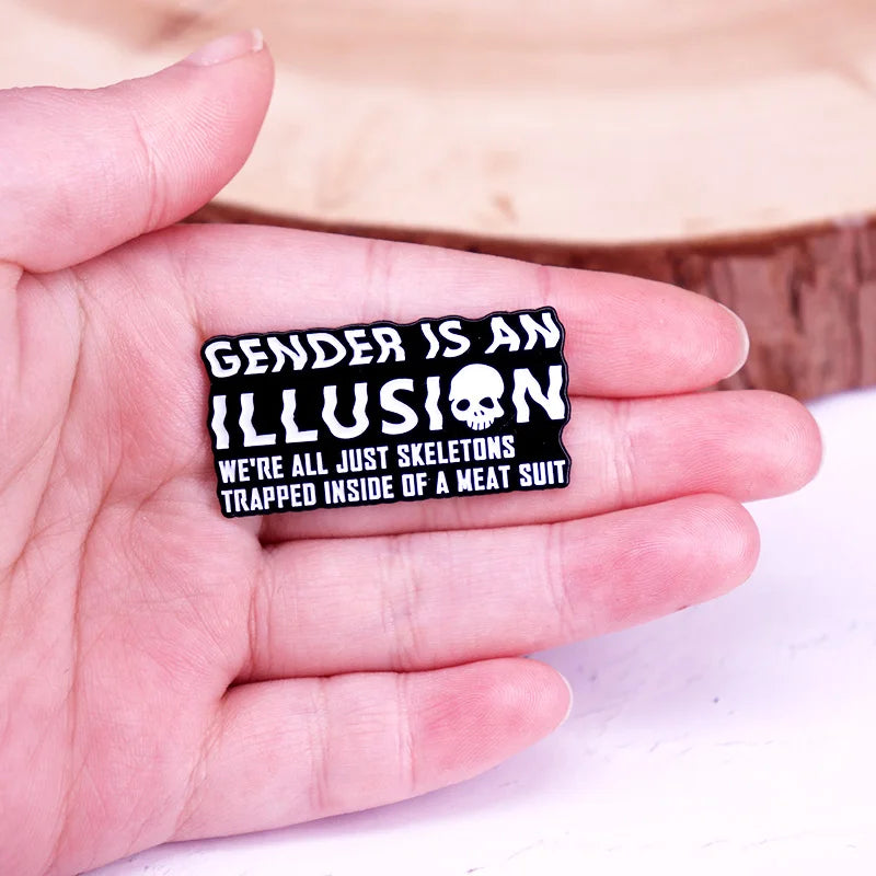 Enamel pin reading “GENDER IS AN ILLUSION” with a skull and skeleton slogan, held in a hand
