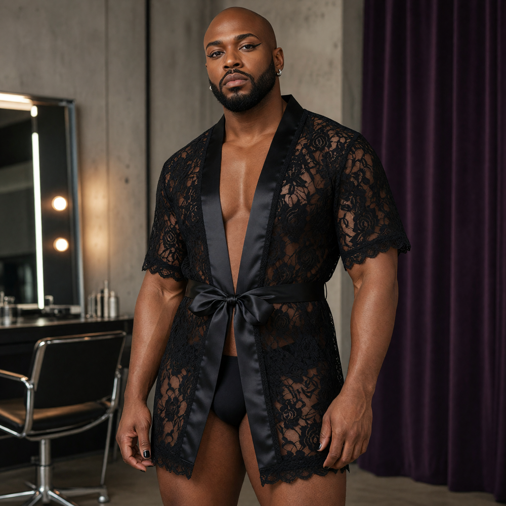 Adult model wearing a short black floral-lace robe with satin edging and waist tie over opaque black underwear in a dressing room.
