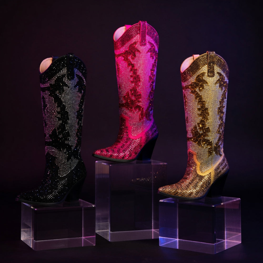 Three rhinestone-covered cowboy boots in black, pink, and gold on clear acrylic blocks