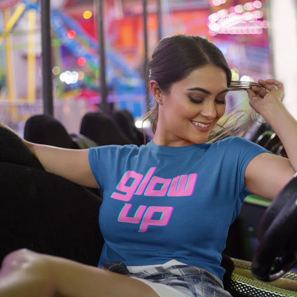 Blue T-shirt with pink and turquoise “glow up” lettering, worn by a seated model