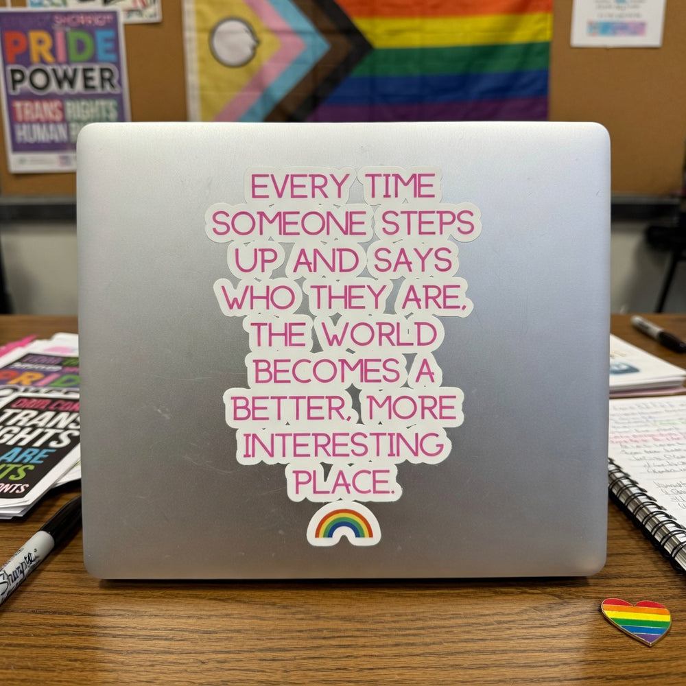 Laptop with "Every Time Someone Steps Up" bubble-free sticker, rainbow, pride flags on corkboard, and notebooks on desk.