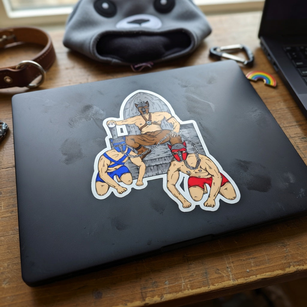 Pup Play Bubble-Free Sticker on a black laptop: three buff dudes in bright wrestling masks. Nearby—dog collar, gray furry hat, rainbow pin.