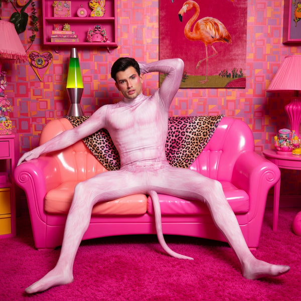 A young man in an Animal Zentai Body Suit With Tail poses confidently on a vibrant pink loveseat, embodying bold queer self-expression and joy in a retro-kitsch styled room.