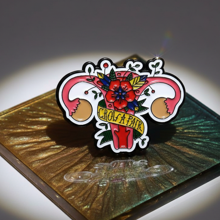 Flowery enamel pin shaped like a pair of flowers with a banner reading “GROW A PAIR”
