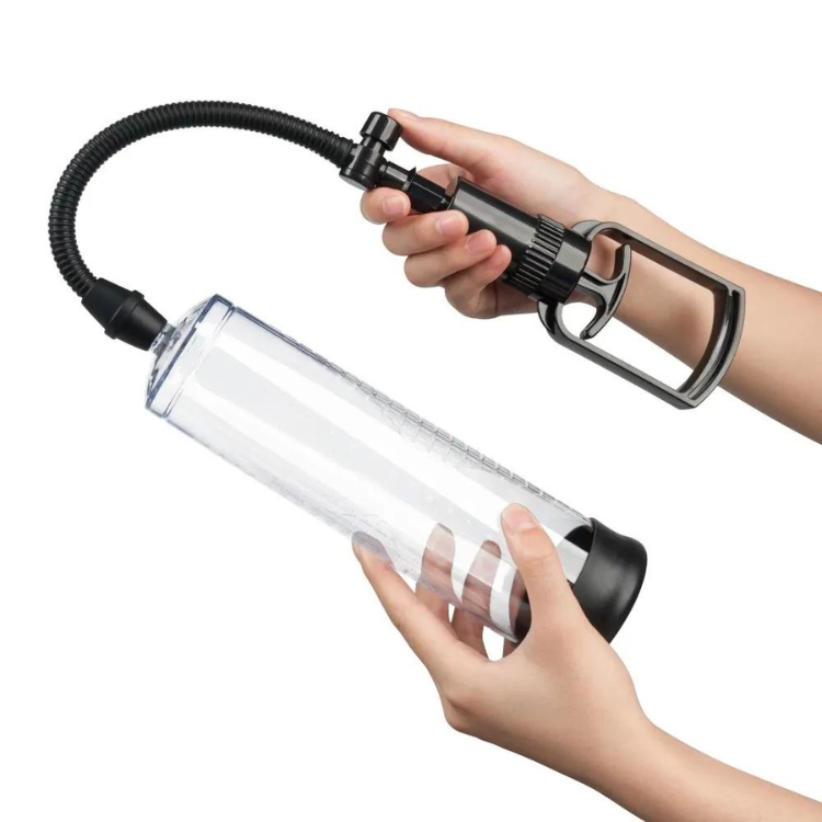 Hands hold a clear penis pump cylinder connected by a hose to a black manual hand pump