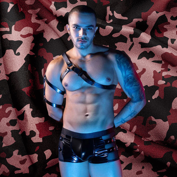 Front-facing model wearing a black leather harness, armbands, and shiny shorts against camouflage