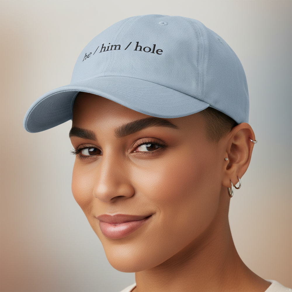 Light blue “He / Him / Hole” cap with black embroidery, worn by a person smiling gently, multiple silver hoops