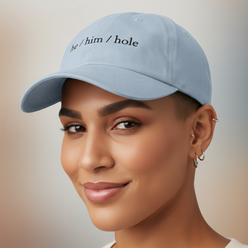 Light blue “He / Him / Hole” cap with black embroidery, worn by a person smiling gently, multiple silver hoops