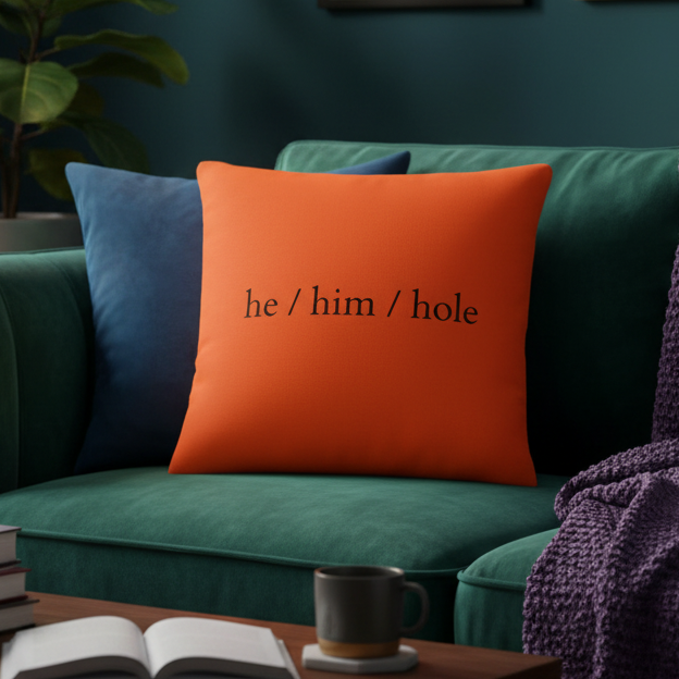 The "He / Him / Hole Pillow" in bold black on bright orange sits on a green sofa with dark blue cushion, books
