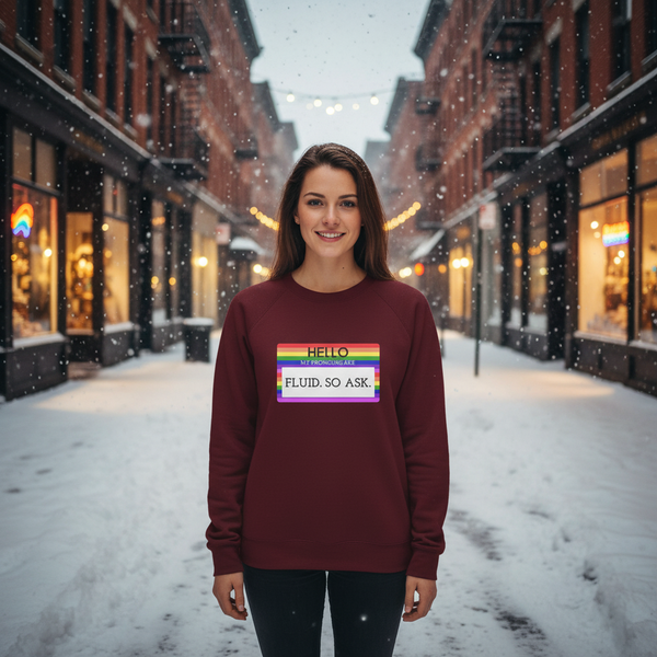 Smiling person in a burgundy sweatshirt with a rainbow-bordered pronoun graphic in a snowy alley