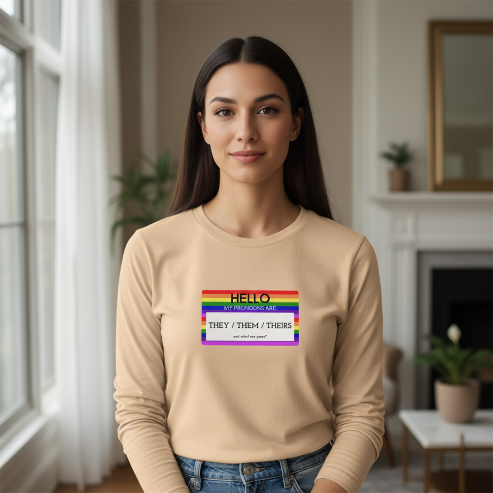 Unisex long sleeve shirt reads "Hello My Pronouns Are They / Them / Theirs." Brown-haired person indoors