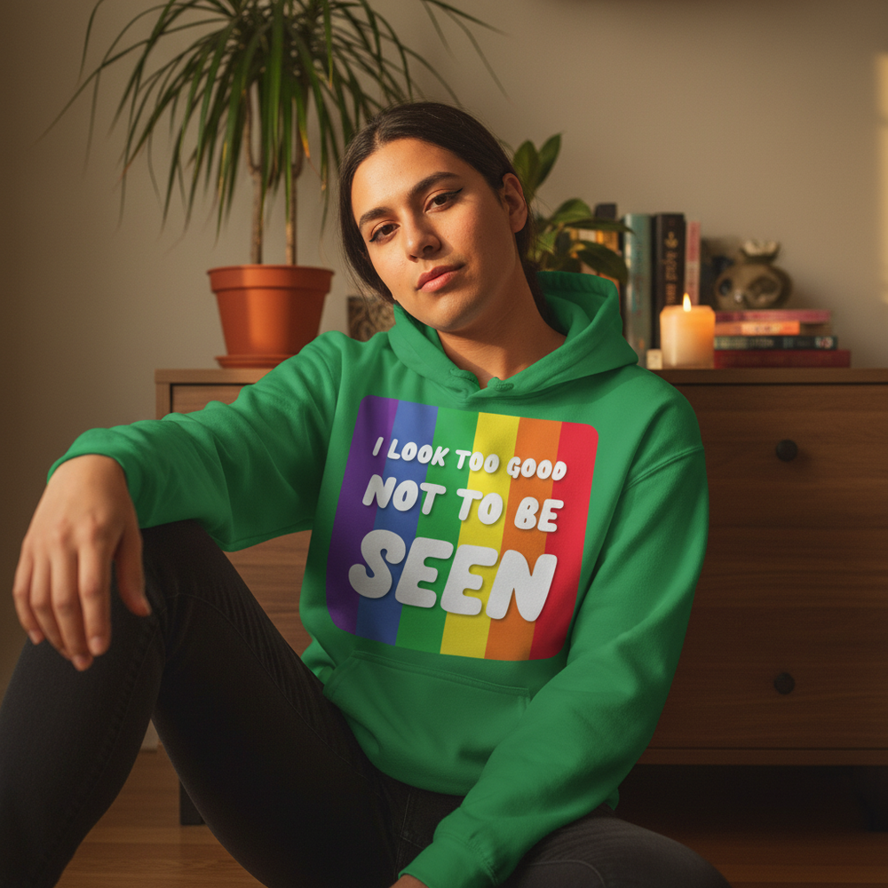 Person seated indoors wearing a green hoodie with rainbow stripes and “I Look Too Good Not To Be Seen” text
