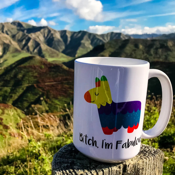 White mug reading “Bitch, I’m Fabulous” beside a colorful llama illustration, outdoors with mountain scenery