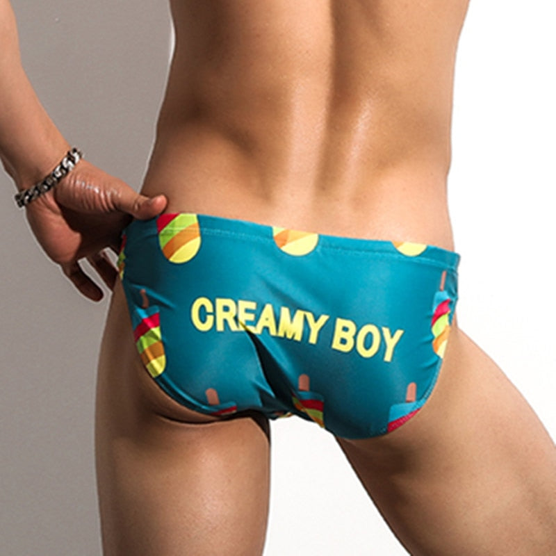 Front view of a model wearing turquoise swim briefs reading “CREAMY BOY,” with ice-cream graphics