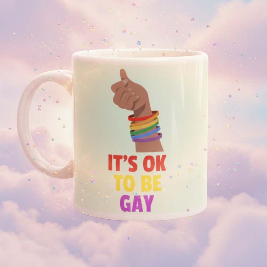 White mug with a raised hand wearing rainbow bracelets and reading “IT’S OK TO BE GAY”