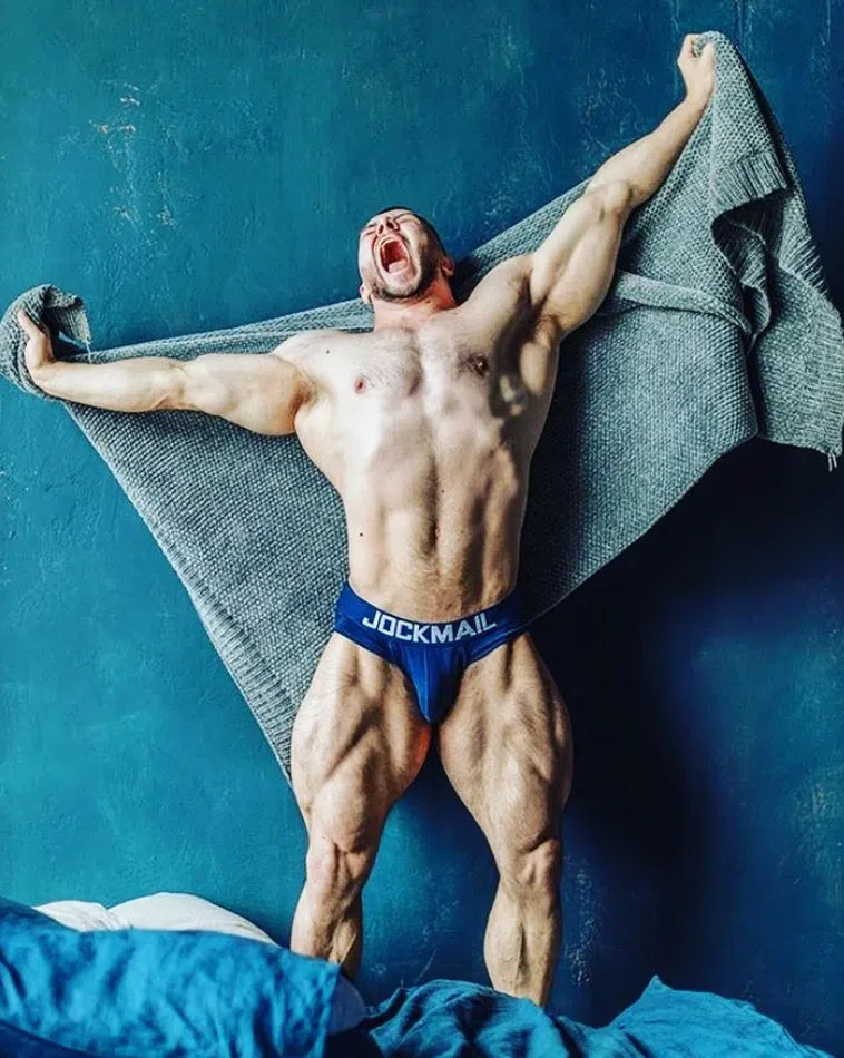 Model wearing blue low-rise Jockmail briefs while holding a gray blanket against a teal wall