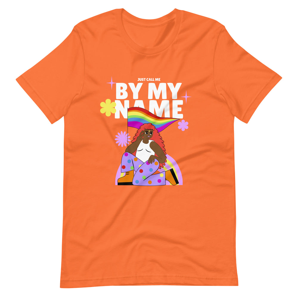 Flat-lay orange T-shirt reading “JUST CALL ME BY MY NAME,” with a rainbow and illustrated woman