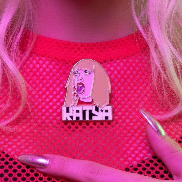 Katya enamel pin with a stylized face and “KATYA” lettering on pink mesh