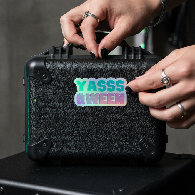 Holographic "YASSS QWEEN" sticker, placed by someone with black nail polish and silver rings, on a textured black hard case.