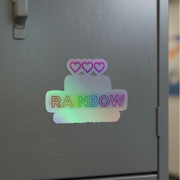 Holographic vinyl sticker showing a rainbow-layer cake, three pink hearts, and bold text "RAINBOW" for your queer sticker stash.