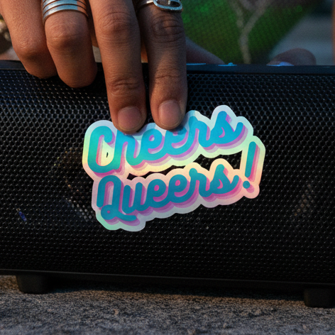 Holographic "Cheers Queers!" sticker shines on a black speaker, lit by sunlight; hand with silver rings adds queer energy.
